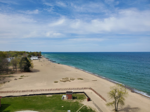 A scenic view of a sandy beach with calm blue waters and a clear sky, lined with trees and distant shoreline.