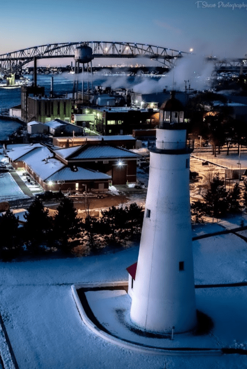 A snowy landscape featuring a lighthouse, industrial buildings, and a bridge in the background, illuminated at dusk.