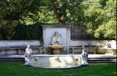 A decorative stone fountain featuring cherubs, surrounded by greenery and a stone wall in a tranquil garden setting.