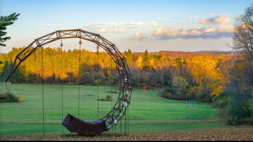 A large metal sculpture resembling a swing in a colorful autumn landscape with trees and a clear sky.