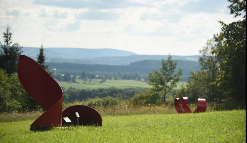 Two abstract red sculptures on a grassy hill, with a scenic view of rolling hills and trees in the background.