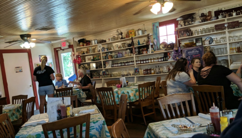A cozy restaurant interior with wooden tables, colorful tablecloths, and shelves filled with dishes and decor.