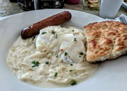 A plate of creamy chicken and noodles with two poached eggs, a sausage link, and a biscuit, garnished with parsley.
