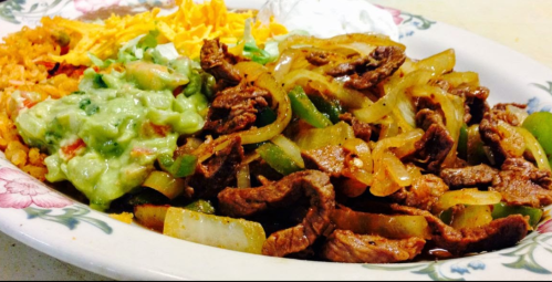 A plate of sizzling beef fajitas with onions and peppers, served with guacamole, rice, cheese, and sour cream.