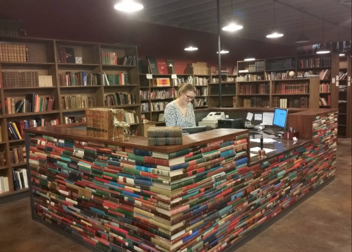 A woman at a reception desk in a library, surrounded by shelves filled with books. The desk features a colorful book design.