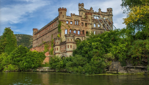 A historic castle partially covered in greenery, situated by a calm river under a blue sky.