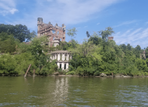A partially overgrown, historic building beside a river, with a blue sky and scattered clouds in the background.