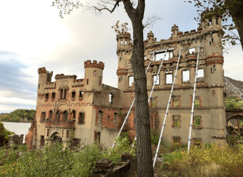 Ruins of a castle-like structure with towers, partially collapsed, surrounded by trees and a river in the background.