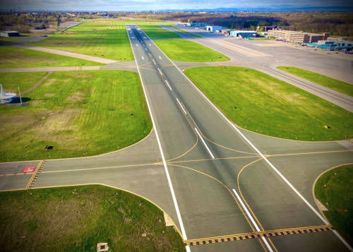Aerial view of an empty airport runway intersecting with taxiways, surrounded by green grass and distant buildings.