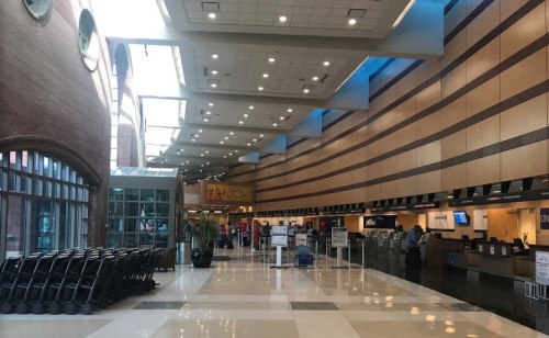 Interior of an airport terminal with check-in counters, seating areas, and luggage carts in a spacious, well-lit environment.
