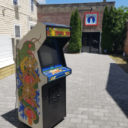 A colorful vintage arcade game stands outside a building with a sign, surrounded by greenery and paved stones.