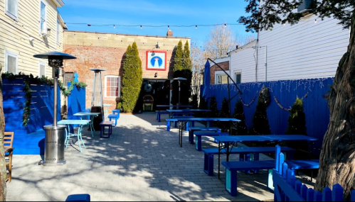 Outdoor patio with blue tables and chairs, string lights, and a mural on the wall, surrounded by greenery.