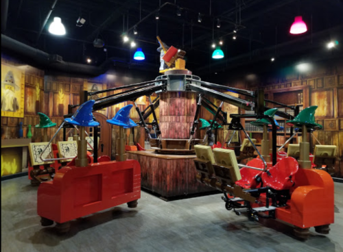 A colorful indoor amusement ride featuring whimsical seats and a central structure with a character holding a wand.