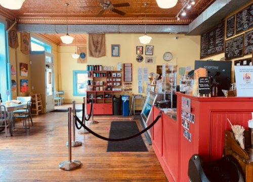 Cozy café interior with wooden floors, red counter, menu board, and seating area, featuring a welcoming atmosphere.