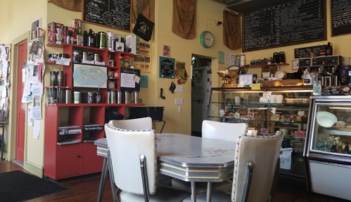 Cozy café interior with a colorful wall, a display case of pastries, and a table with chairs in a warm atmosphere.