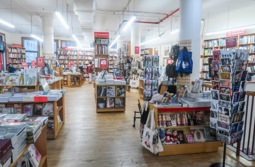 A cozy bookstore interior with wooden floors, shelves filled with books, and displays of magazines and merchandise.