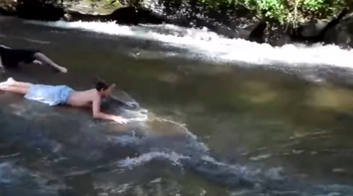 Two people play in a shallow stream, one lying on their stomach while the other swims nearby.