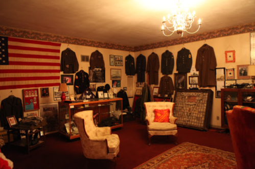 A vintage room displaying military uniforms, an American flag, and various historical artifacts, with two armchairs and a chandelier.