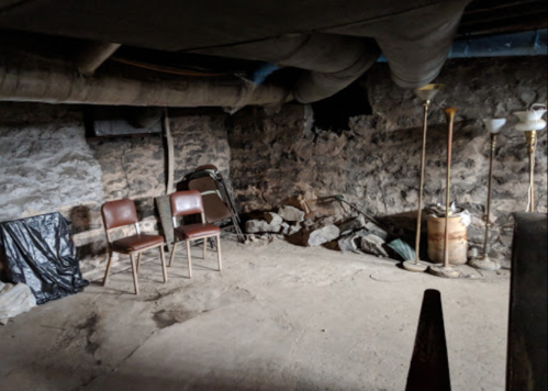 A dimly lit basement with stone walls, two chairs, a pile of rocks, and various lamps scattered around.