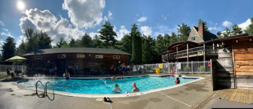 A sunny pool scene with people swimming and relaxing, surrounded by trees and a rustic building in the background.