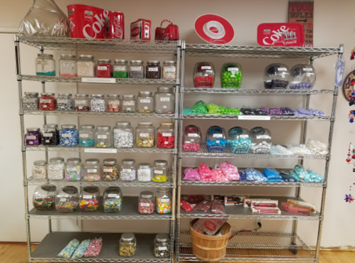 A colorful display of jars filled with various candies on metal shelves, with a Coca-Cola theme in the background.