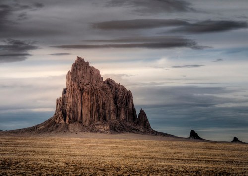 A dramatic rock formation rises from a barren landscape under a cloudy sky, showcasing natural beauty and rugged terrain.