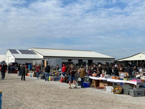 A bustling outdoor market with people browsing tables filled with various items, set against a cloudy sky.