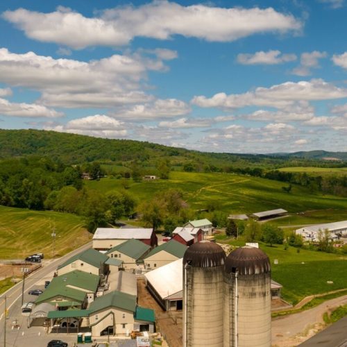 Aerial view of a farm with silos, buildings, and green fields under a partly cloudy sky.
