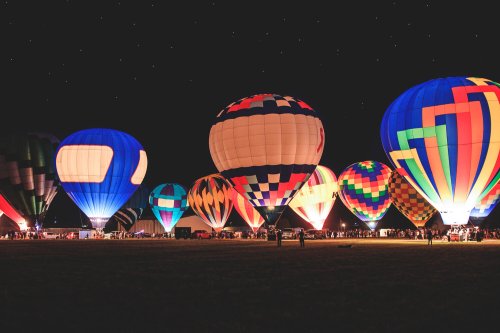 Colorful hot air balloons illuminated at night, set against a starry sky with a crowd gathered below.