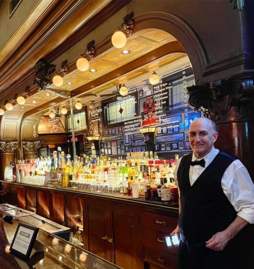 A bartender in a tuxedo stands at a vintage bar, surrounded by shelves of various liquor bottles and framed photos.