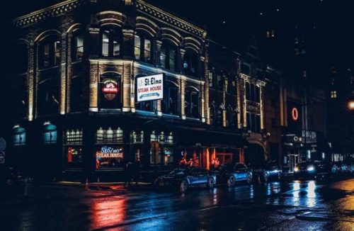 A lit-up steakhouse at night, with reflections on wet pavement and cars parked outside.