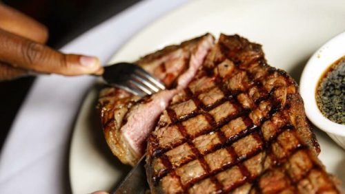 A close-up of a grilled steak being sliced with a fork and knife, served with a small dish of seasoning.