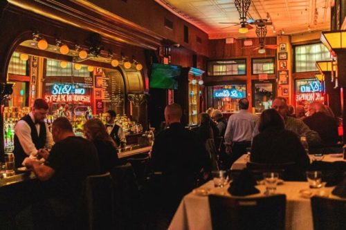 A lively restaurant bar scene with patrons dining and bartenders serving drinks in a warmly lit atmosphere.