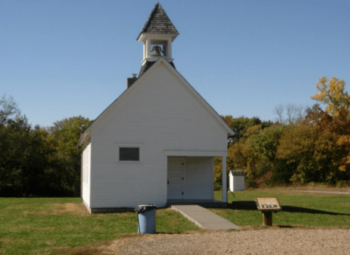 A small white church with a bell tower, surrounded by green trees and a gravel path leading to a sign and trash can.