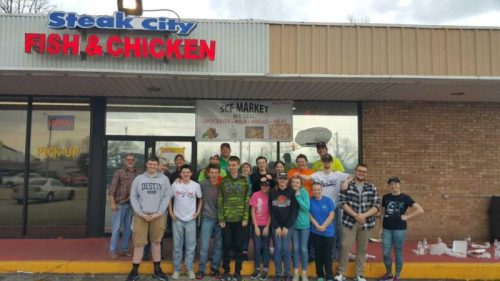A group of people poses outside a restaurant called Steak City Fish & Chicken, with a sign for SCF Market above.