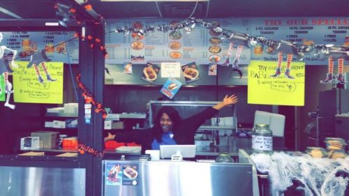 A cheerful employee stands behind a counter in a vibrant restaurant, surrounded by colorful signs and decorations.