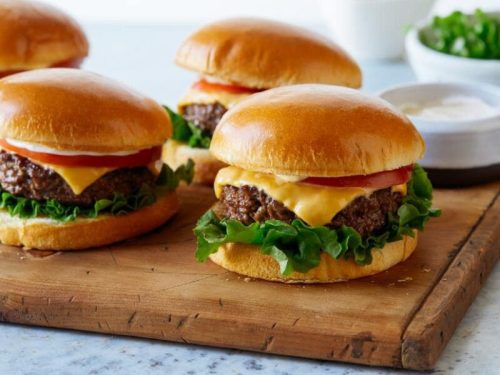 Three juicy cheeseburgers with lettuce, tomato, and soft buns, served on a wooden cutting board.