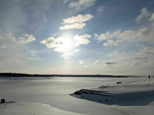 A serene winter landscape with a frozen lake, snow-covered ground, and a bright sun partially obscured by clouds.