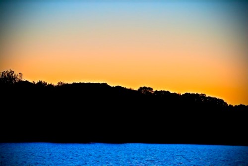Silhouette of trees against a gradient sunset sky, reflecting on calm water below.