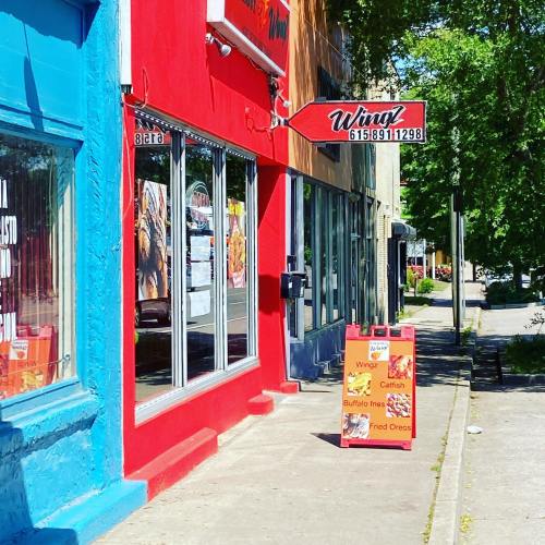 Colorful storefronts with a sign for "Wings!" and a sandwich board advertising menu items on a sunny day.