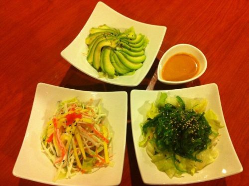 Three small bowls of salad: one with avocado slices, one with mixed vegetables, and one with seaweed, plus a dipping sauce.