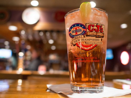A glass of iced tea with a lime wedge, featuring logos from C.J.'s and Bones restaurants, on a wooden bar.