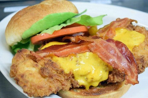 A close-up of a chicken sandwich with crispy chicken, melted cheese, bacon, lettuce, and tomato on a toasted bun.