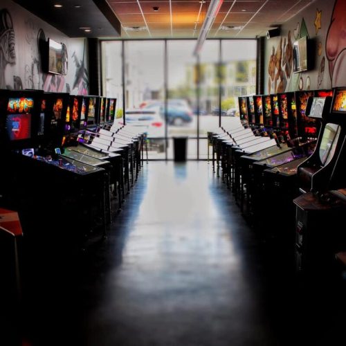A row of arcade machines in a brightly lit room, with large windows in the background.