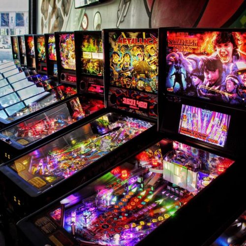 A row of colorful pinball machines featuring themes from Metallica and Stranger Things, illuminated with vibrant lights.