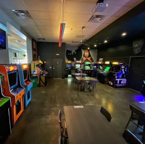 A vibrant arcade with classic game machines, tables, and a dark wall featuring a mural.