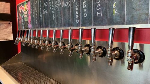 A row of shiny beer taps mounted on a wall, with a chalkboard menu above listing various drink options.