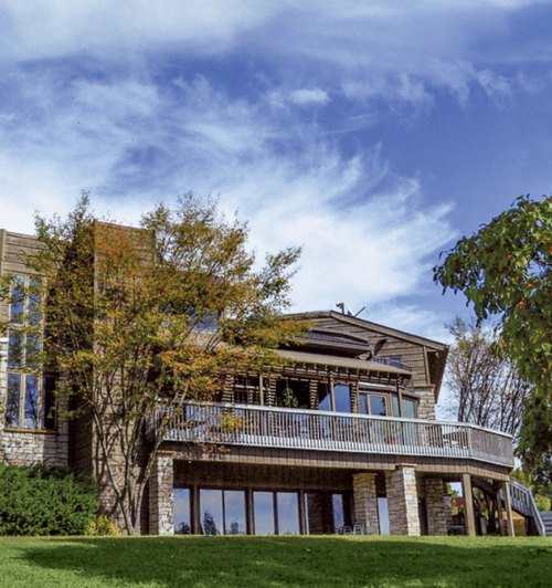 A modern house with large windows and a deck, surrounded by trees under a blue sky with scattered clouds.