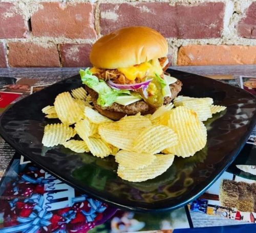A delicious burger with lettuce, cheese, and pickles, served with a side of crispy potato chips on a black plate.