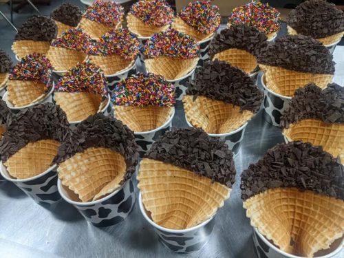 A variety of ice cream cones with chocolate and rainbow sprinkles, arranged in cups on a silver surface.